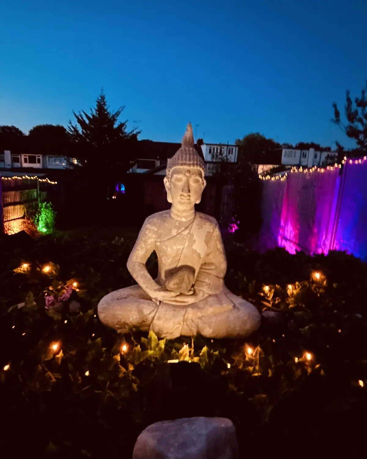 Her garden at dusk: a stone Buddha among candles, coloured lights along the fence